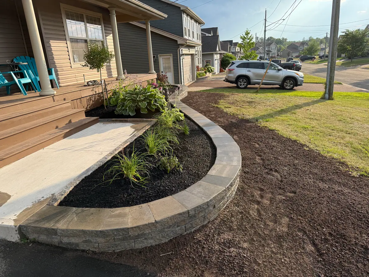 Curved segmental block raised bed along a front porch, newly planted and mulched, with graded soil ready for lawn